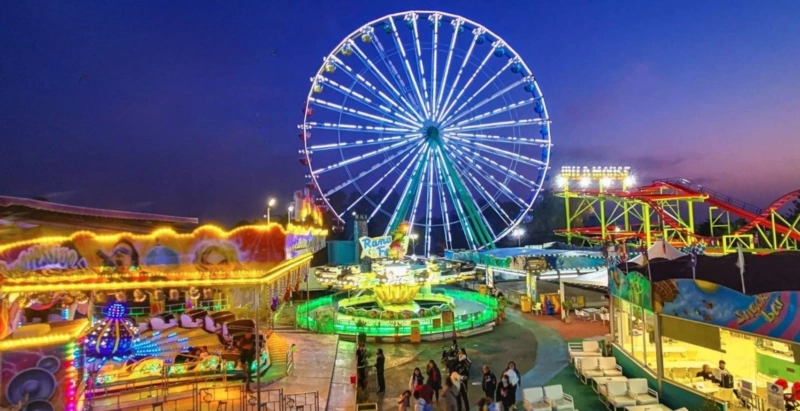 Colourful illuminated Ferris wheel and rides at Parko Paliatso amusement park at dusk in Ayia Napa, Cyprus.