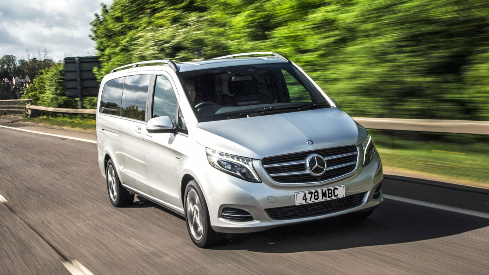 A silver Mercedes-Benz passenger van drives along a tree-lined road in daylight. The image captures the vehicle in motion, highlighting its spacious design and suitability for family travel, airport transfers, or group transportation