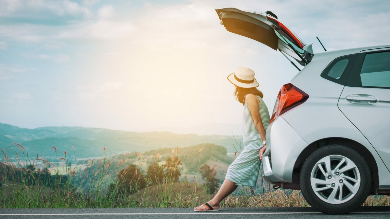 A woman wearing a sun hat sits on the open tailgate of a white hatchback, enjoying a scenic mountain view at sunset. Parked by the roadside, the car overlooks rolling hills and open countryside, evoking a sense of freedom, travel, and relaxation.