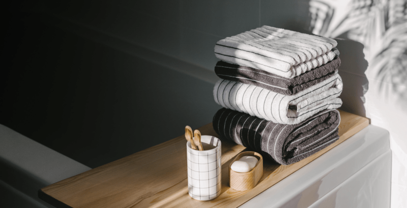A neatly arranged stack of folded towels sits on a wooden bath tray beside a bathtub, accompanied by a soap dish and a cup holding bamboo toothbrushes. Soft natural light creates gentle shadows, giving the modern bathroom a calm, spa-like atmosphere.