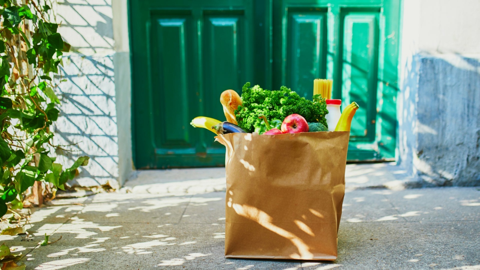A reusable grocery bag filled with fresh groceries, including leafy greens, bread, bananas, and apples, sits on a doorstep in front of a green front door, suggesting a home grocery delivery.