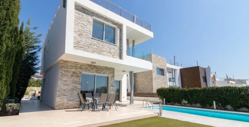 A contemporary holiday villa with striking stone-clad walls, spacious balconies, and a private lap pool. The outdoor terrace features a dining area overlooking the landscaped garden, creating an ideal space for relaxing and entertaining during a Cyprus getaway.