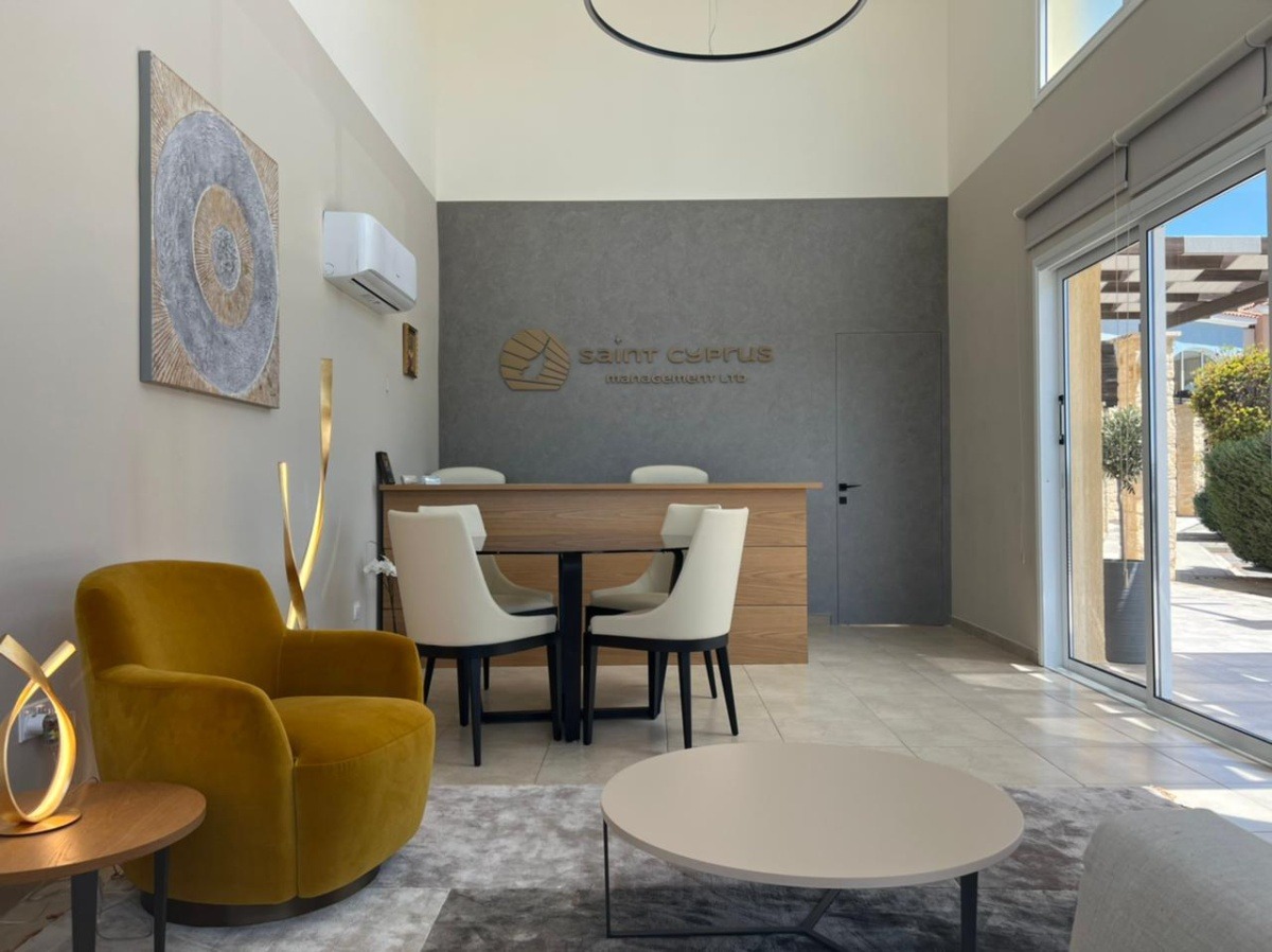 Modern office reception area with a high ceiling, a wooden reception desk, white guest chairs, a mustard-yellow lounge chair, contemporary lighting, and large glass doors letting in natural light. The Saint Cyprus Management Ltd logo is displayed on the feature wall behind the reception desk.