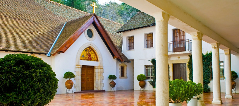 White chapel with a red-tiled roof beside the arcaded buildings of Trooditissa Monastery, surrounded by greenery in the Troodos Mountains of Cyprus.