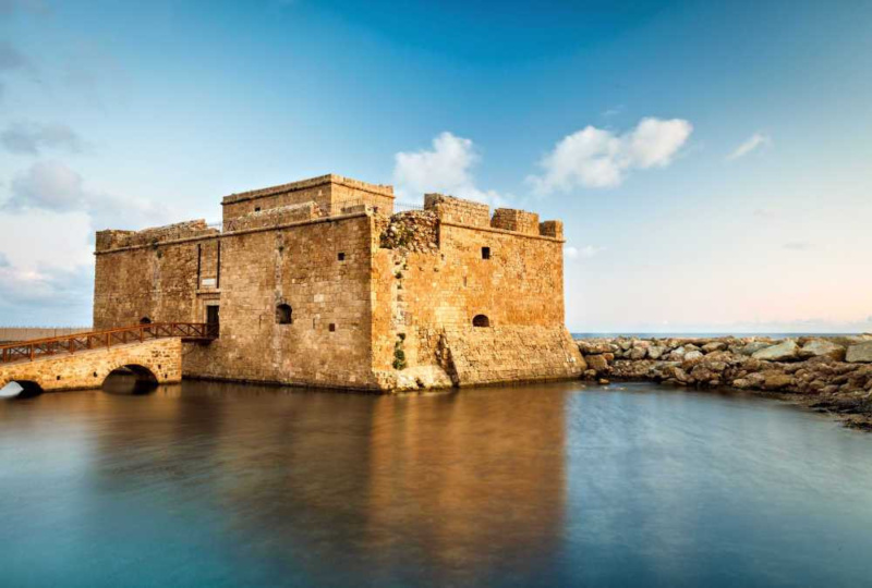 The medieval stone Paphos Harbour Castle reflected in the calm harbour water under a blue sky in Cyprus.