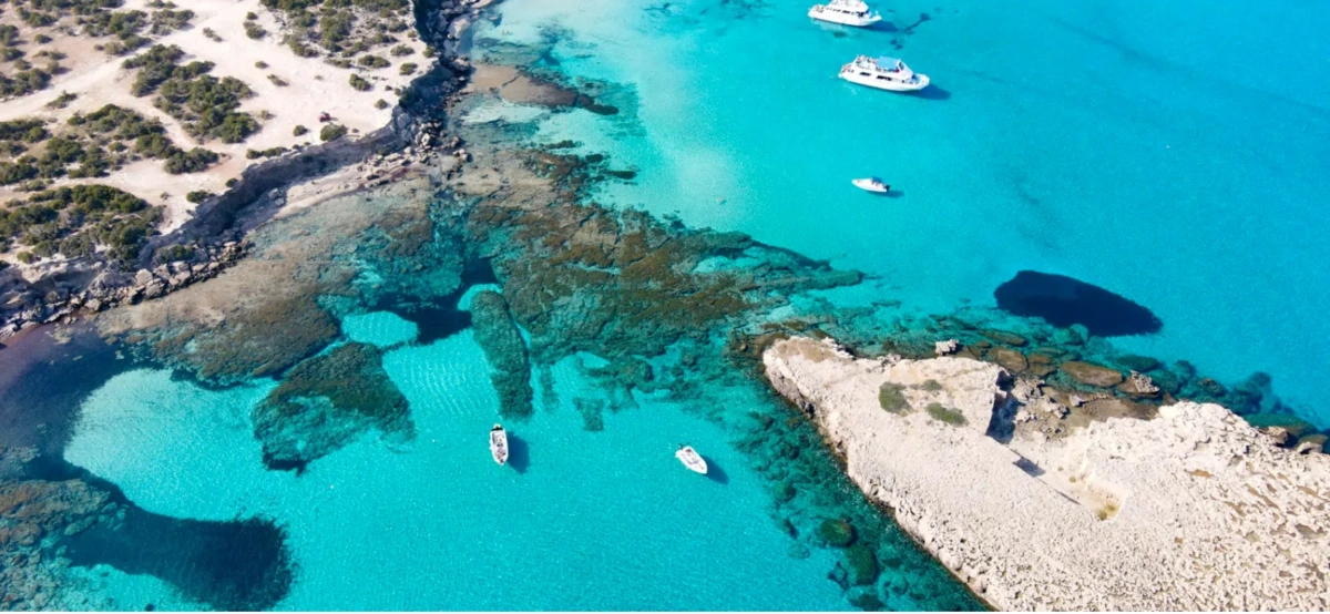 Aerial view of the Blue Lagoon on the Akamas peninsula in Cyprus, with boats floating on crystal-clear turquoise water beside a rocky coastline.