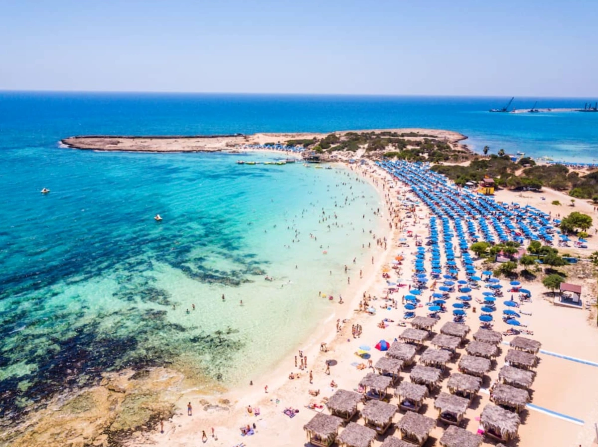 A long sandy beach lined with rows of blue sun umbrellas and sunbeds beside calm turquoise sea in Cyprus.