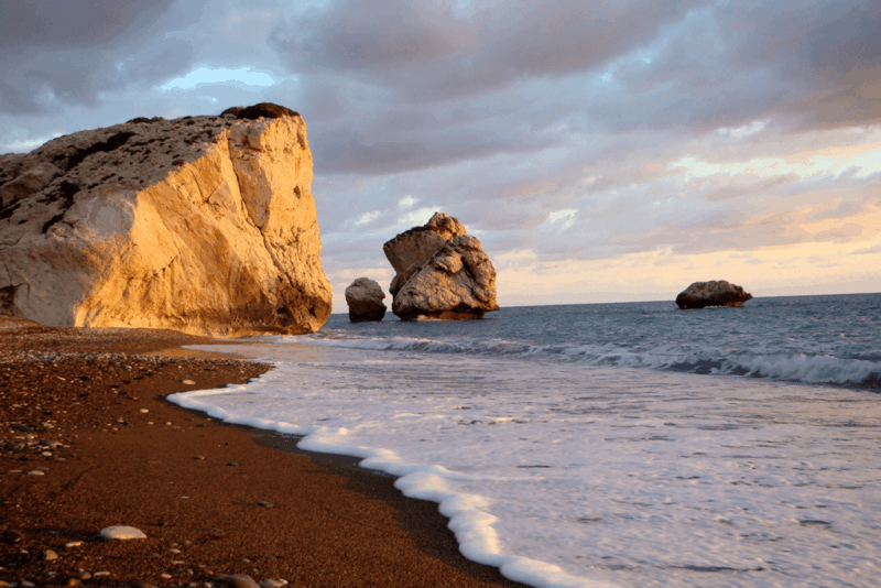 Golden sunlight illuminates the iconic white limestone sea stacks along a sandy beach as gentle waves wash onto the shore. Dramatic clouds overhead and the calm Mediterranean Sea create a striking coastal landscape, showcasing one of Cyprus's most famous natural landmarks.