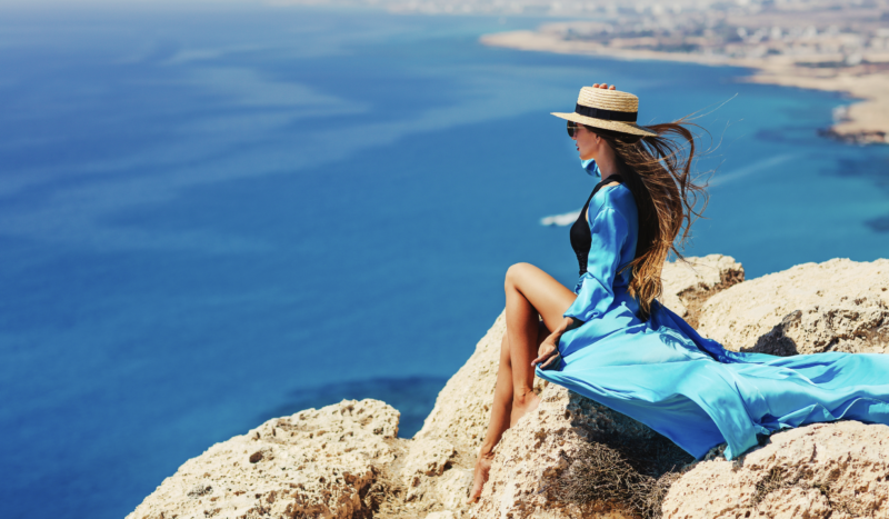 A woman in a flowing blue dress and straw hat sits on a rocky cliff overlooking a stunning turquoise coastline. With the wind blowing through her hair, she gazes out over the sea and distant shoreline, capturing the beauty and tranquility of a luxury coastal destination.
