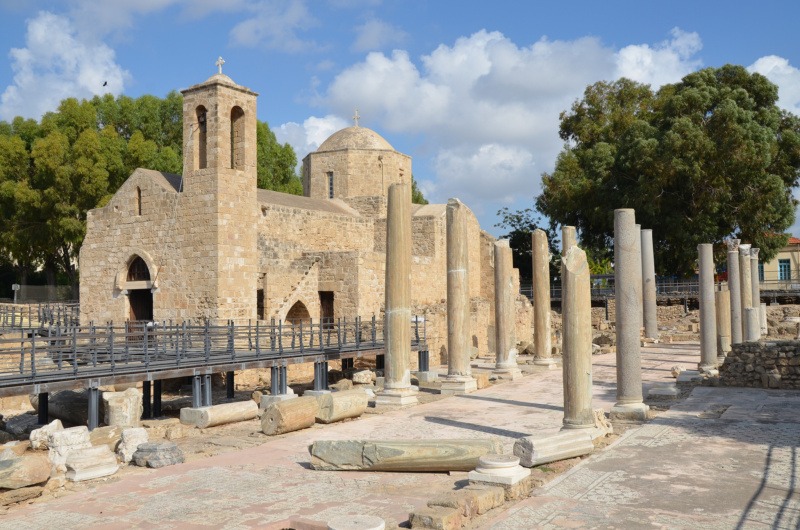 The historic Church of Panagia Chrysopolitissa stands beside the ancient archaeological site in Paphos, Cyprus. Surrounded by Roman columns and archaeological ruins, the well-preserved stone church reflects the island's rich cultural and religious heritage under a bright blue sky.