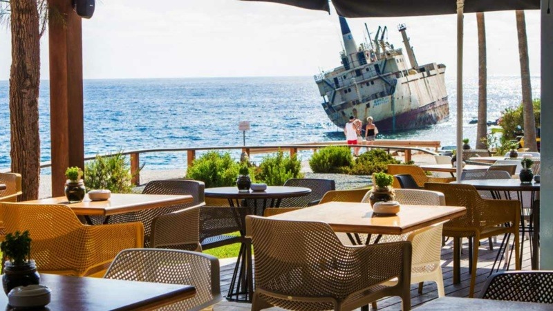 Seaside restaurant terrace of Oniro by the Sea with tables set for dining and a view over the blue Mediterranean and a shipwreck near Paphos, Cyprus.