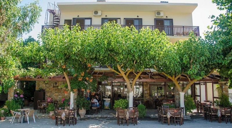 A charming countryside guesthouse with a shaded outdoor restaurant terrace sits beneath lush green trees. Stone walls, wooden tables, and a peaceful garden setting create a welcoming atmosphere, ideal for dining and relaxing in a traditional Cypriot village.